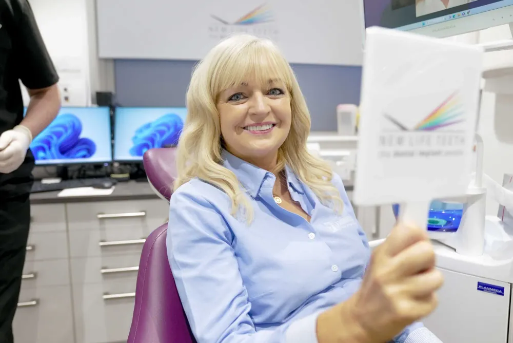 Smiling New Life Teeth patient relaxing in the treatment chair in a branded clinic, holding a hand mirror